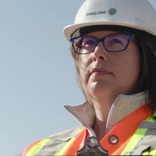 Portrait of Lindsay Freckleton wearing a hard hat and high-visibility safety vest stands outdoors against a clear sky, with trees visible in the background. 