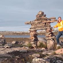photo of Lydia charbonneau wearing a high-visibility safety jacket stands beside a stone inukshuk in a rocky, open landscape near water. Low buildings and industrial structures are visible in the distance under a wide sky.