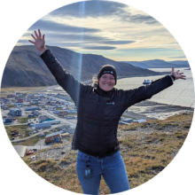Lydia Charbonneau standing on a hill with arms raised, overlooking a northern coastal town and mountains under a cloudy sky.