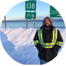 Person wearing a high‑visibility winter safety jacket standing beside snowy roadside signs marking Route 138 and the end of the road, with an ocean backdrop under a clear sky.