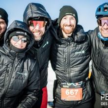 Englobe Team wearing Englobe-branded winter coats standing together on a snowy course at the Snow Pentathlon, with tall buildings and event structures in the background.