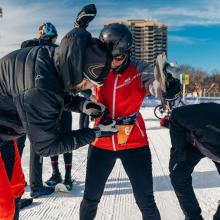 Englobe participants assisting a teammate in the transition zone of the Snow Pentathlon, adjusting gear and preparing for the next event on a snowy course.