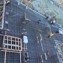 Overhead view of a construction site showing workers installing dense steel rebar and formwork for a reinforced concrete foundation slab, with excavation walls and temporary wooden structures surrounding the area.