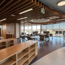 Interior view of victoria high school library featuring wooden ceiling slats, built‑in bookshelves, study tables, computer workstations, and large floor‑to‑ceiling windows bringing in natural light.