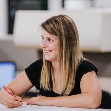 Woman sitting at a desk writing with a red pen, she is looking off towards the left