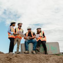 A diverse group of four construction professionals in safety gear review blueprints and take notes on a construction site, emphasizing teamwork and collaboration under a bright sky.