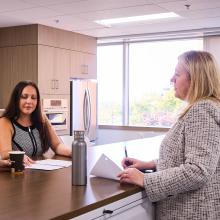Two professional women collaborate in an office, with one taking notes and the other reviewing documents, highlighting a casual yet productive work environment.