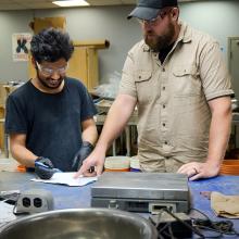 Two technicians in safety glasses collaborate over a document on a blue workbench in a laboratory, surrounded by equipment, showcasing a focus on innovation and precision.