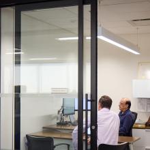 A professional office setting featuring two men engaged in discussion, a desk with computer equipment, and a framed "Professional Engineer" certificate on the wall.