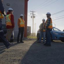 A small group of workers wearing hard hats and high-visibility safety vests stand and walk outside an industrial building on a gravel surface. Parked vehicles, utility poles, and overhead power lines are visible nearby, with low-angle sunlight casting long shadows across the ground.