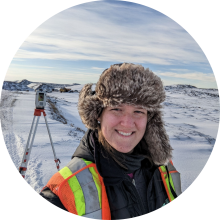 A person wearing a winter safety jacket with high‑visibility stripes and a fur‑lined hat stands in a snowy, open landscape, with a surveying instrument on a tripod set up behind them and snow‑covered hills under a partly cloudy sky.