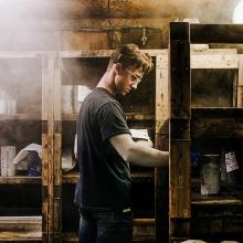 A person stands indoors beside wooden shelving units, handling small labeled containers in a workshop or storage area. Dust or steam is visible in the air, and the space appears industrial, with rough wood surfaces and materials stacked on shelves.