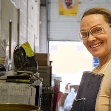A person wearing a protective apron stands in an industrial workshop beside a workbench with metal containers, tools, and paperwork. The space includes concrete walls, shelving, a small fan, and an open garage-style door letting in natural light, suggesting a hands-on work environment.