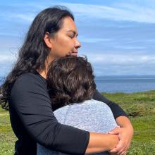 Indigenous mother and child stand outdoors on a grassy shoreline, embracing each other while facing a calm body of water. The scene is framed by open sky and distant land across the water, creating a quiet, natural setting.
