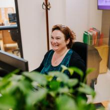 A woman sits at a desk in a bright office workspace, working at a computer. A leafy plant is visible in the foreground, with a coat rack, filing folders, and office furniture in the background, suggesting a professional administrative setting.