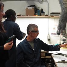 Natacha Sénéchal working with colleagues inside a laboratory, examining cylindrical material samples placed on a work surface. Shelving, containers, and ventilation equipment are visible in the workspace.