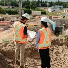 Two young englobe's employees wearing hard hats and high-visibility safety vests stand on a dirt embankment at an active construction site, reviewing a set of plans. Excavation work, concrete forms, and construction materials are visible below, with buildings, trees, and utility lines in the background.