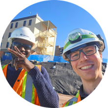 Two construction workers wearing safety vests and hard hats at an outdoor worksite with buildings and equipment in the background.