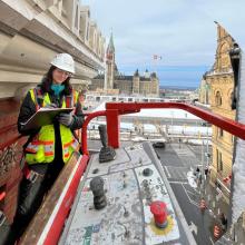 Kylie Bennett conducting a building inspection from an aerial lift beside an ornate brick façade with Parliament Hill in the background.