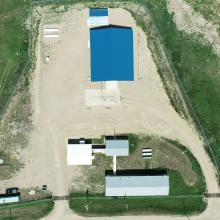Aerial view of a rural emergency water treatment facility showing multiple buildings with blue roofs, access roads, fenced perimeter, and surrounding grassland.