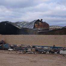 Fire‑damaged industrial building with collapsed roof sections, exposed structural elements, and debris scattered around the exterior under an overcast sky.
