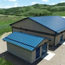 Exterior view of an emergency water treatment building with blue metal roofing, gravel access areas, and rolling green hills in the background.