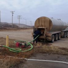 Tanker truck connected to hoses transferring water at a rural supply point, with fuel tanks, gravel ground, and utility infrastructure nearby.