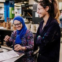Two colleagues collaborate in an industrial workshop, one seated and reviewing technical documents at a workstation while the other stands nearby, with machinery, tools, and materials visible around them.