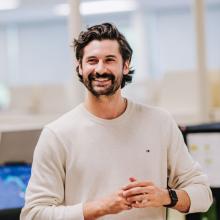 A professional standing in a modern office environment, wearing a light-colored sweater and smartwatch, with hands loosely clasped while speaking or presenting, and computer monitors visible in the background.