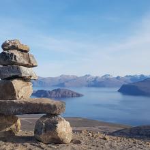 Scenic coastal landscape featuring a stacked rock cairn in the foreground, overlooking a calm blue fjord with scattered islands and distant snow-capped mountains under a clear blue sky.