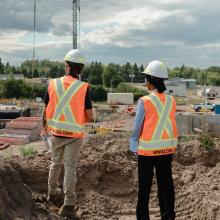 Two construction workers in orange safety vests and white hard hats overseeing an active construction site with excavated foundations, a crane, and industrial buildings in the background.
