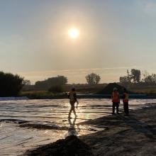 Three construction workers in safety gear at a site during sunrise or sunset, with a tarp-covered area, a large pipe, and a hazy industrial backdrop illuminated by warm sunlight.