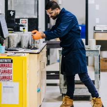 A person wearing a blue protective coat, orange gloves, and work boots stands at a laboratory workbench, handling containers beside a computer workstation. The setting is a laboratory environment with cabinets, safety equipment, carts, and a yellow flammable materials storage unit visible nearby.