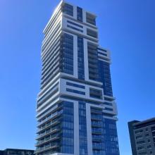 Contemporary high-rise residential building with staggered balconies and large glass façades, photographed against a clear blue sky in an urban setting.