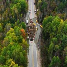 Aerial view of a two-lane rural road undergoing repairs, with construction equipment, traffic cones, and temporary barriers set within a dense forested landscape.