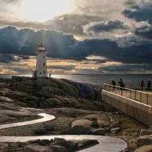 Coastal lighthouse on rocky terrain beside a winding boardwalk, overlooking the ocean under dramatic cloud formations with sunlight breaking through.