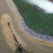 Aerial view of a curved river channel with flowing water bordered by riprap stone armoring, alongside a gravel access road and stabilized shoreline in a managed natural landscape.