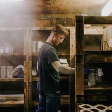 Laboratory interior showing a person working at wooden shelving units, with containers and materials stored in a dimly lit, dusty environment.