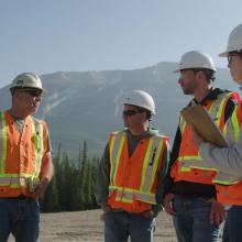 Group of professionals wearing hard hats and high-visibility vests discussing a construction site with mountains in the background.
