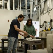 Two coworkers working together at a table in an industrial workspace, reviewing material samples and tools.