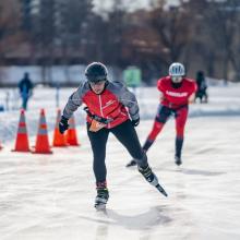 Genevieve Lemieux racing on an outdoor ice track during the Snow Pentathlon, with orange cones marking the course and trees and buildings in the background.