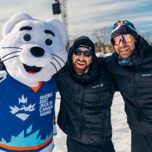 Mascot in a blue Québec 2027 Jeux du Canada Games jersey standing beside two participants in Englobe jackets at the Snow Pentathlon.