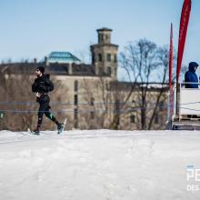 William Carmichael running on a snowy course at the Snow Pentathlon, with spectators watching from a raised platform and historic buildings in the background.