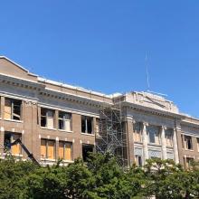 Exterior view of victoria High school building undergoing construction, with scaffolding, protective blue tarps, boarded windows, and restoration work visible under a clear blue sky.