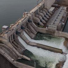 Aerial view of a large hydroelectric dam with concrete spillways releasing water into a river, surrounded by construction areas and access roads.