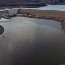 Aerial view of a hydroelectric dam and reservoir under construction, showing spillway structures, earthworks, and water channels surrounded by a natural landscape.