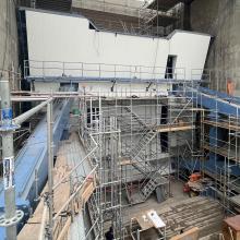 Exterior view of a massive concrete hydroelectric dam under construction, showing spillway outlets, scaffolding, heavy lifting equipment, and active construction work at the base of the structure.