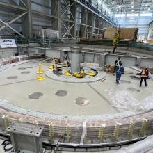 Interior view of a hydroelectric power station turbine installation, featuring a large circular turbine assembly inside an industrial facility with construction equipment and safety barriers.